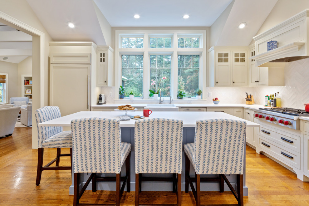 Example of a large beach style l-shaped medium tone wood floor and brown floor kitchen pantry design in Boston with a farmhouse sink, shaker cabinets, white cabinets, quartz countertops, white backsplash, ceramic backsplash, stainless steel appliances, an island and white countertops
