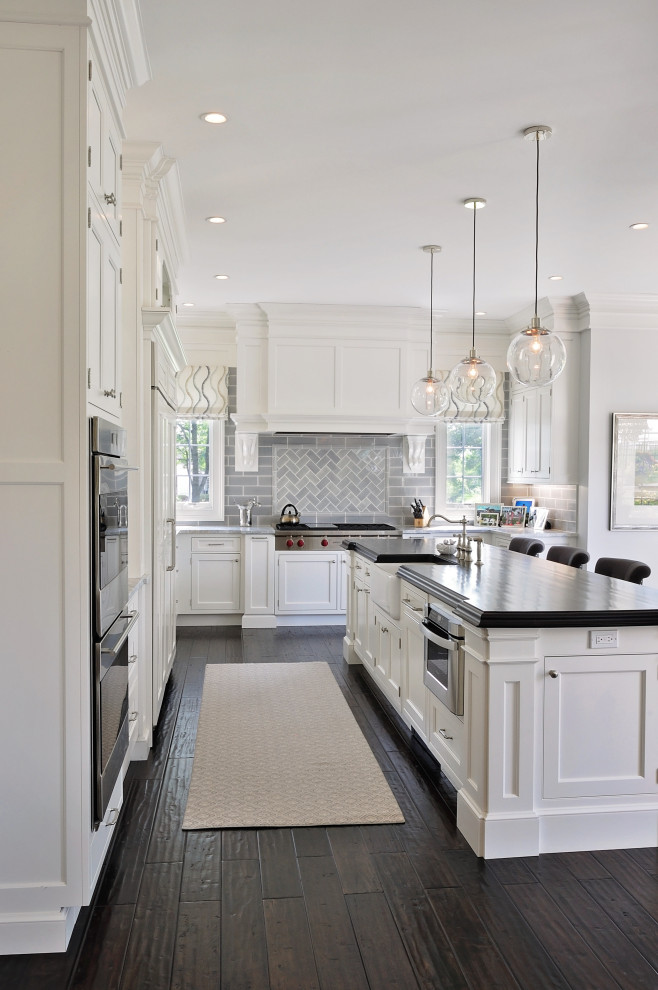 This is an example of a mid-sized traditional l-shaped eat-in kitchen in Philadelphia with a farmhouse sink, recessed-panel cabinets, white cabinets, granite benchtops, grey splashback, subway tile splashback, panelled appliances, dark hardwood floors, with island, brown floor and grey benchtop.