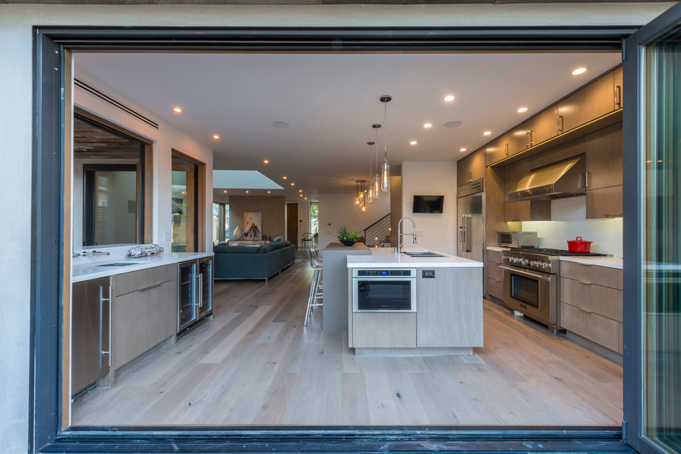 This is an example of a mid-sized contemporary l-shaped open plan kitchen in Los Angeles with a double-bowl sink, flat-panel cabinets, light wood cabinets, solid surface benchtops, white splashback, glass sheet splashback, stainless steel appliances, medium hardwood floors, with island and brown floor.