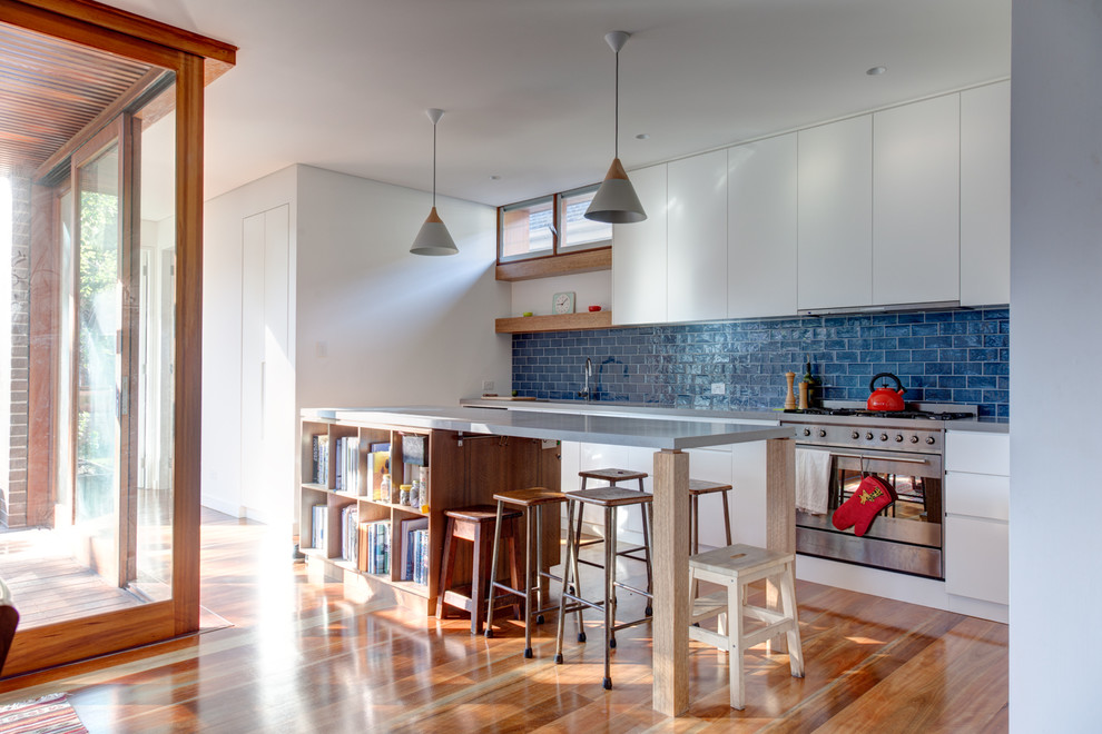 Contemporary galley open plan kitchen in Sydney with flat-panel cabinets, white cabinets, blue splashback, stainless steel appliances, medium hardwood floors, with island, brown floor and grey benchtop.