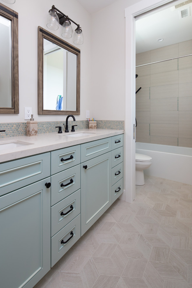 This is an example of a mid-sized beach style kids bathroom in San Diego with beaded inset cabinets, blue cabinets, an alcove tub, a shower/bathtub combo, a two-piece toilet, beige tile, porcelain tile, grey walls, porcelain floors, an undermount sink, engineered quartz benchtops, beige floor, a shower curtain and white benchtops.