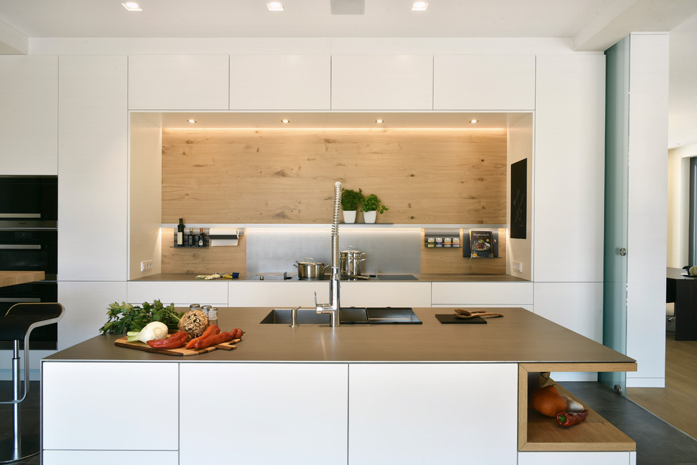 Mid-sized contemporary galley separate kitchen in Frankfurt with an integrated sink, flat-panel cabinets, white cabinets, brown splashback, timber splashback, black appliances, multiple islands, grey floor and grey benchtop.