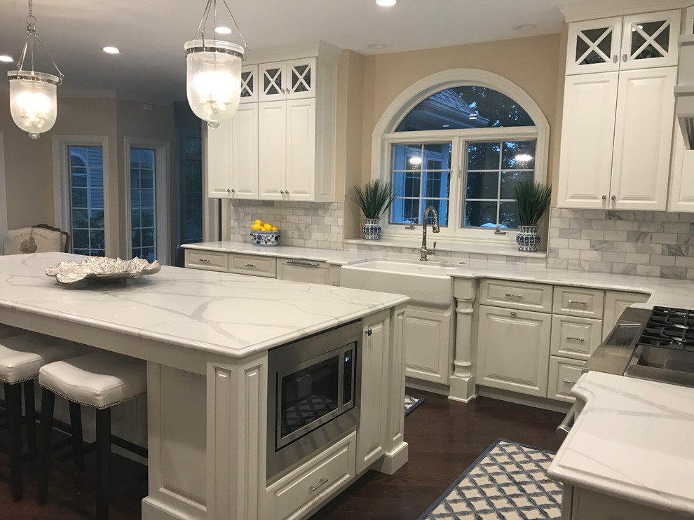 Kitchen - l-shaped dark wood floor kitchen idea with a farmhouse sink, raised-panel cabinets, white cabinets, quartz countertops, gray backsplash, marble backsplash, stainless steel appliances and an island