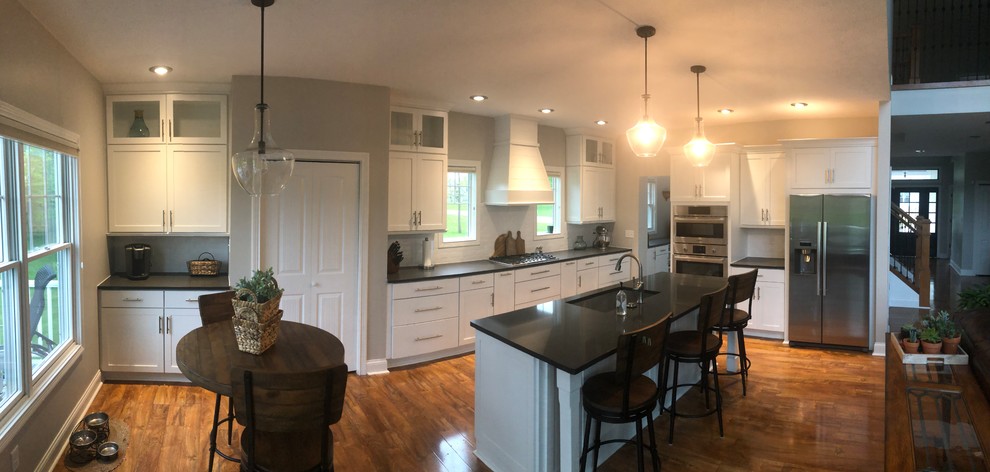 Example of a laminate floor and brown floor kitchen design with a single-bowl sink, shaker cabinets, white cabinets, quartzite countertops, white backsplash, subway tile backsplash, stainless steel appliances, an island and gray countertops