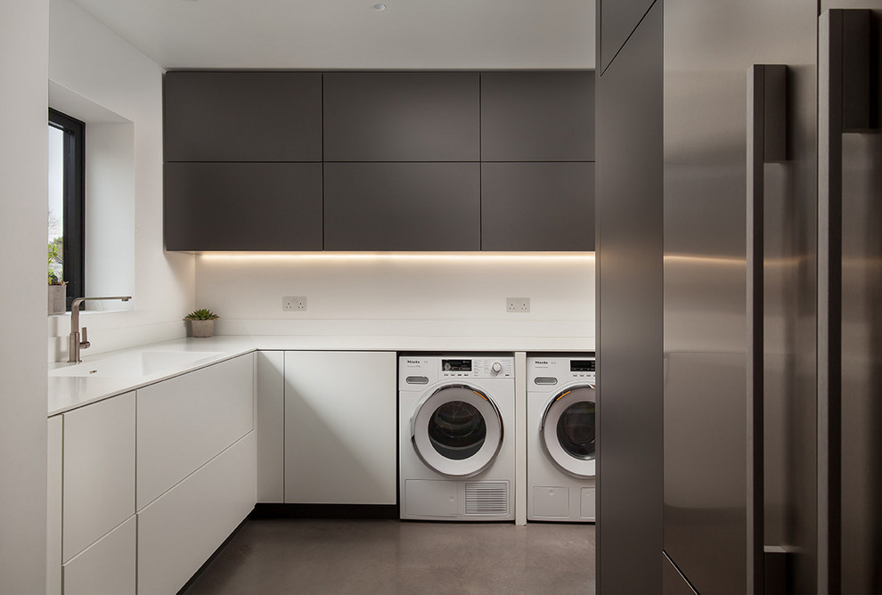 Photo of a modern l-shaped utility room in Cornwall with an integrated sink, flat-panel cabinets, grey cabinets, white walls, concrete floors, a side-by-side washer and dryer, grey floor and white benchtop.
