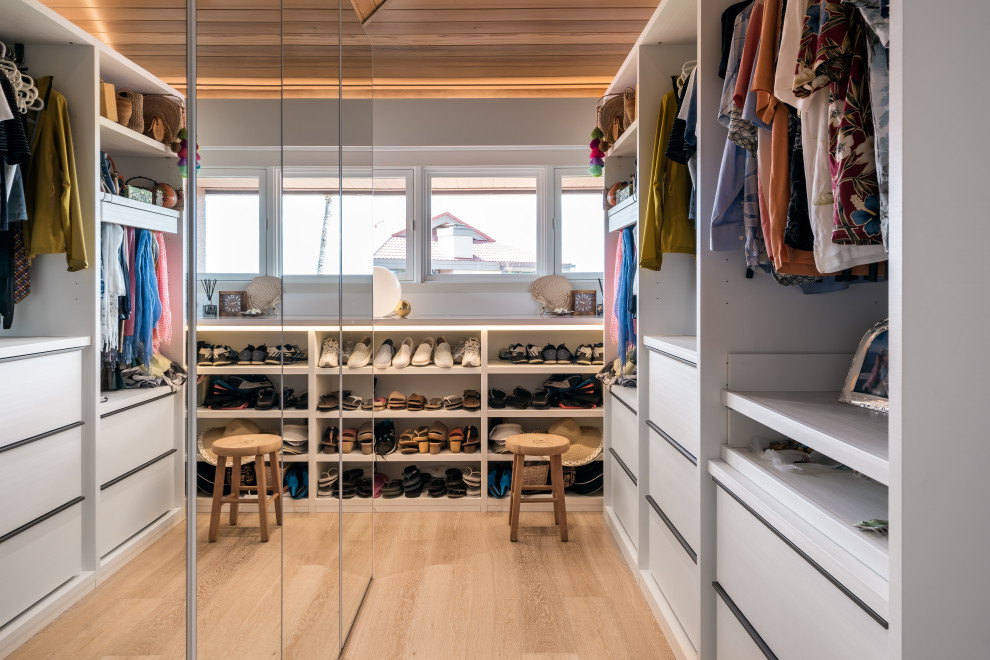Photo of a small modern gender-neutral walk-in wardrobe in Hawaii with glass-front cabinets, white cabinets, light hardwood floors, beige floor and vaulted.