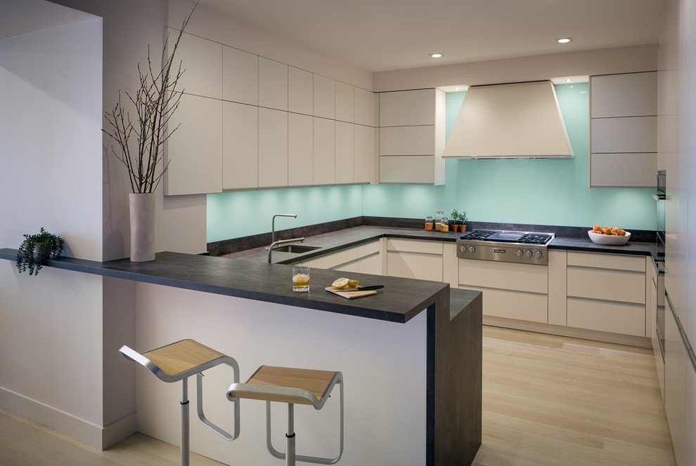 Photo of a mid-sized contemporary u-shaped kitchen in San Francisco with flat-panel cabinets, beige cabinets, solid surface benchtops, blue splashback, glass sheet splashback, stainless steel appliances, light hardwood floors, a peninsula and a single-bowl sink.