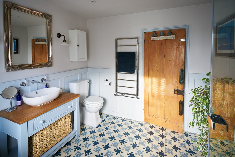 Photo of a large traditional master bathroom in Sussex with raised-panel cabinets, blue cabinets, a freestanding tub, an open shower, a two-piece toilet, gray tile, grey walls, cement tiles, a trough sink, wood benchtops, multi-coloured floor and a hinged shower door.