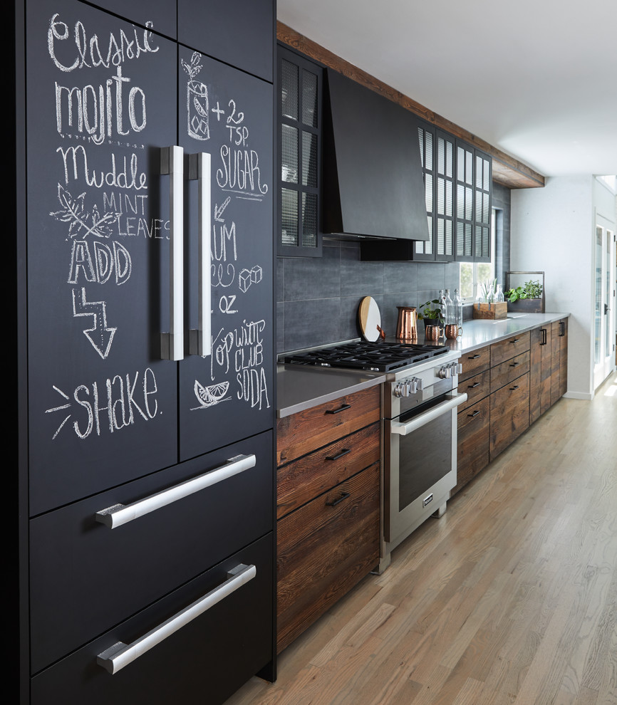This is an example of a large industrial galley eat-in kitchen in New York with an undermount sink, flat-panel cabinets, dark wood cabinets, grey splashback, black appliances, light hardwood floors, with island, beige floor and grey benchtop.