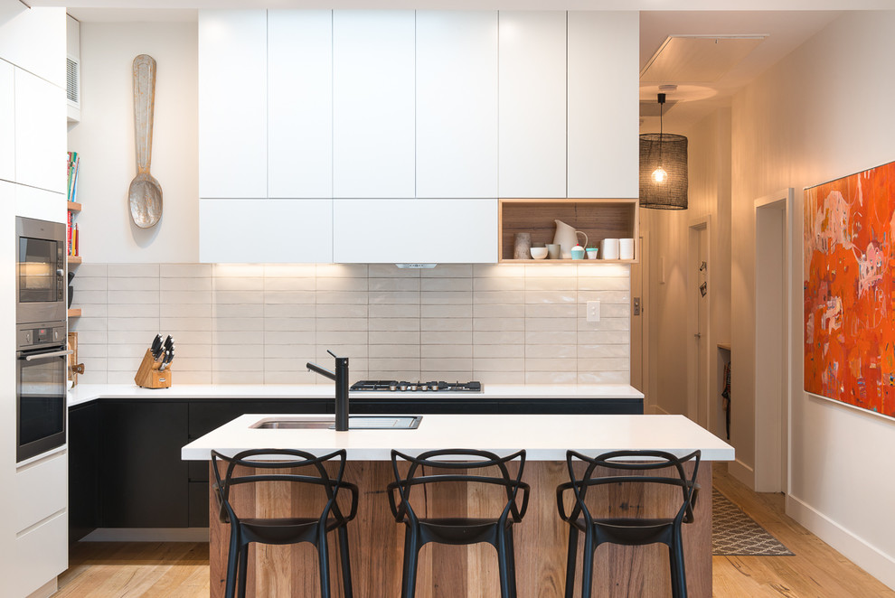 Small contemporary l-shaped open plan kitchen in Melbourne with a drop-in sink, flat-panel cabinets, white splashback, porcelain splashback, stainless steel appliances, light hardwood floors, beige floor and multiple islands.