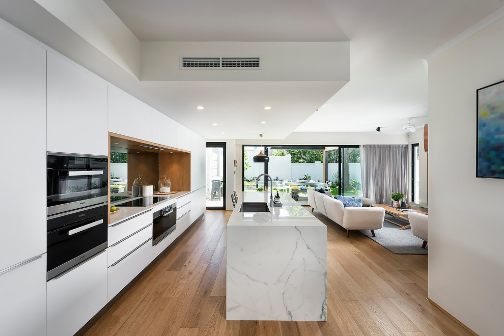 This is an example of a large scandinavian galley open plan kitchen in Perth with a single-bowl sink, flat-panel cabinets, white cabinets, quartz benchtops, metallic splashback, mirror splashback, black appliances, light hardwood floors, with island, brown floor and white benchtop.