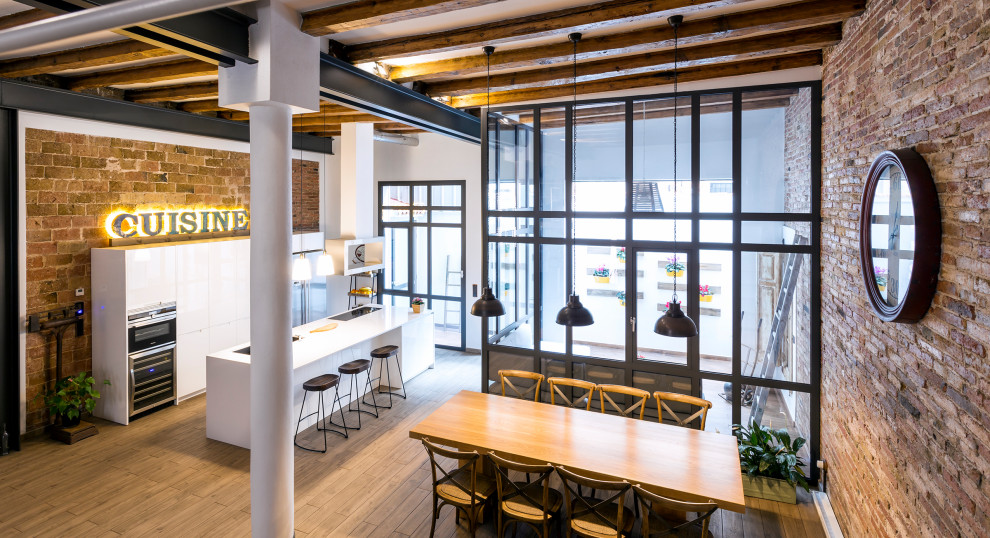 Large industrial single-wall open plan kitchen in Barcelona with an undermount sink, flat-panel cabinets, white cabinets, laminate benchtops, stainless steel appliances, porcelain floors, with island, brown floor, white benchtop and exposed beam.