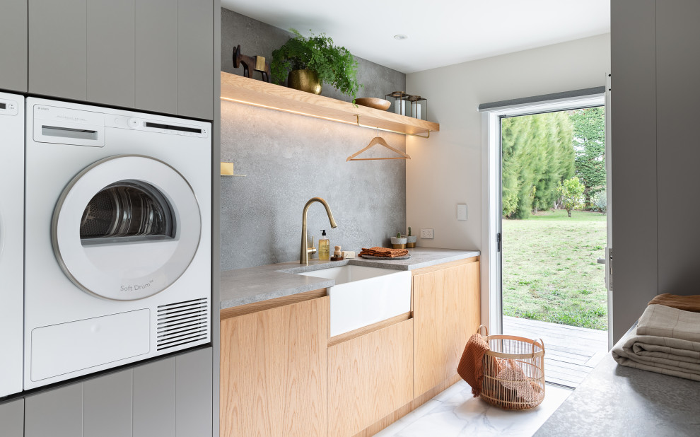 Photo of a mid-sized contemporary galley dedicated laundry room in Auckland with a farmhouse sink, flat-panel cabinets, light wood cabinets, quartz benchtops, grey splashback, porcelain splashback, white walls, porcelain floors, a side-by-side washer and dryer, white floor and grey benchtop.