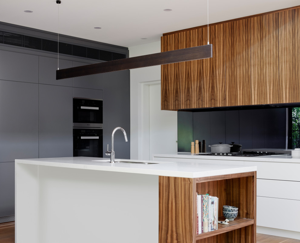 Design ideas for a mid-sized contemporary galley open plan kitchen in Melbourne with an undermount sink, flat-panel cabinets, white cabinets, solid surface benchtops, black splashback, glass sheet splashback, panelled appliances, medium hardwood floors, with island and white benchtop.