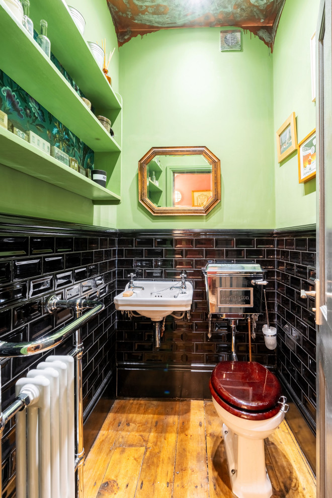 Photo of a bohemian cloakroom in Manchester with a two-piece toilet, black tiles, metro tiles, green walls, medium hardwood flooring, a wall-mounted sink and brown floors.