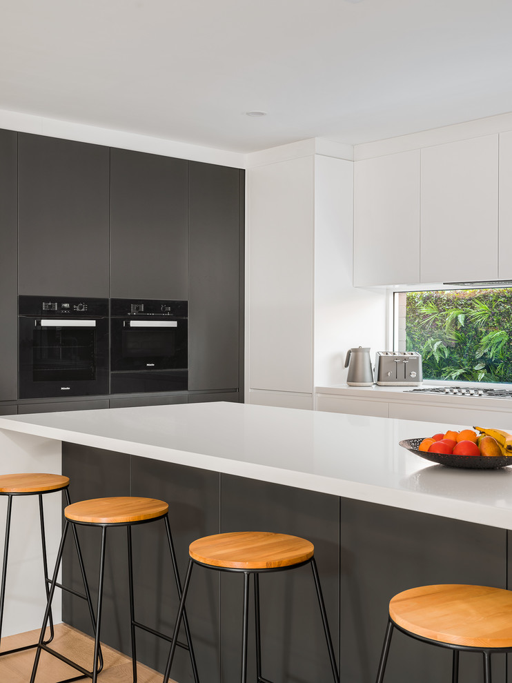 Inspiration for a large modern l-shaped open plan kitchen in Sydney with a double-bowl sink, open cabinets, dark wood cabinets, solid surface benchtops, white splashback, window splashback, black appliances, light hardwood floors, with island, brown floor and white benchtop.