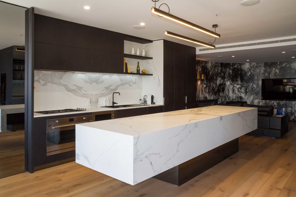 Photo of a mid-sized modern galley open plan kitchen in Melbourne with a drop-in sink, flat-panel cabinets, black cabinets, tile benchtops, white splashback, porcelain splashback, stainless steel appliances, medium hardwood floors, with island, brown floor and white benchtop.