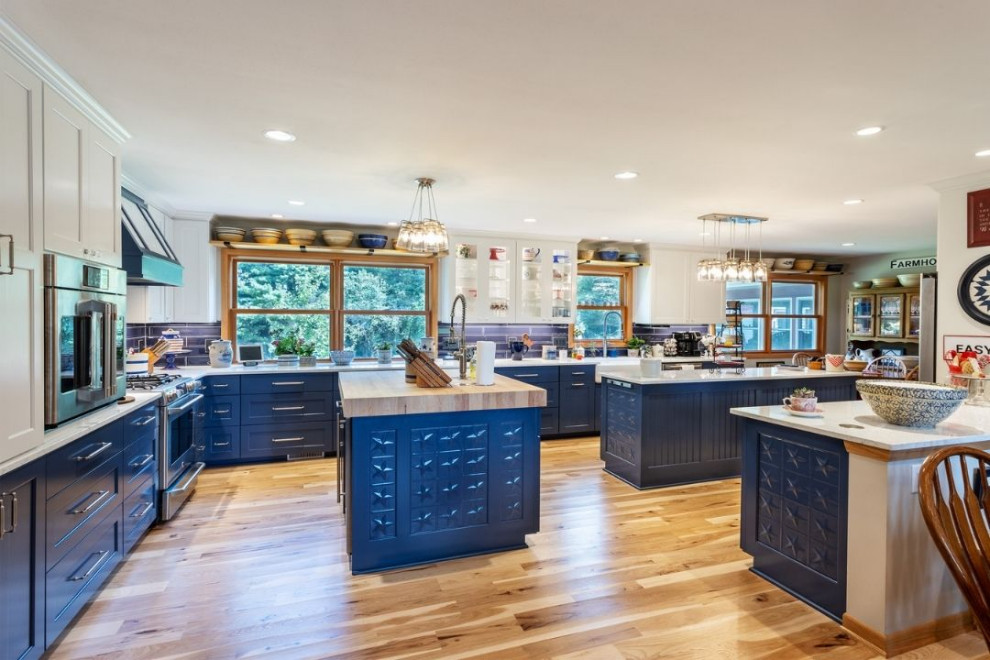 This is an example of a large eclectic l-shaped open plan kitchen in Other with a farmhouse sink, shaker cabinets, blue cabinets, quartz benchtops, blue splashback, subway tile splashback, stainless steel appliances, light hardwood floors, multiple islands, beige floor and white benchtop.