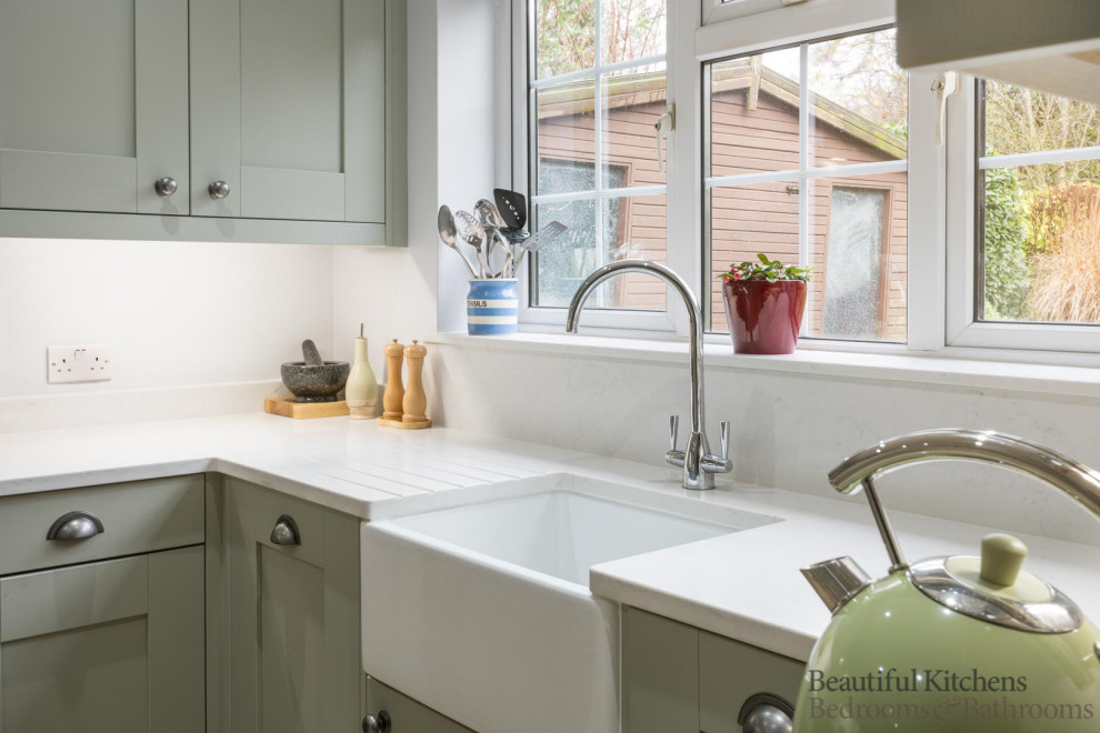 This is an example of a mid-sized country l-shaped eat-in kitchen in Surrey with a farmhouse sink, shaker cabinets, green cabinets, quartzite benchtops, white splashback, stone slab splashback, black appliances, laminate floors, a peninsula, brown floor, white benchtop and vaulted.