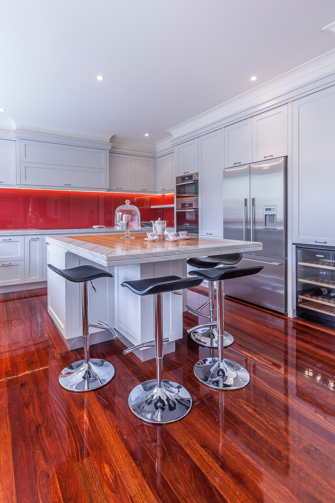 Photo of a large traditional u-shaped eat-in kitchen in Auckland with an undermount sink, recessed-panel cabinets, granite benchtops, red splashback, glass sheet splashback, stainless steel appliances, dark hardwood floors, with island, brown floor and white cabinets.