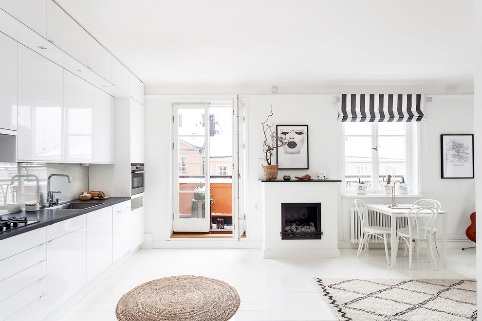 This is an example of a large scandinavian single-wall open plan kitchen in Stockholm with a single-bowl sink, flat-panel cabinets, white cabinets, grey splashback, glass sheet splashback, painted wood floors, laminate benchtops and white appliances.