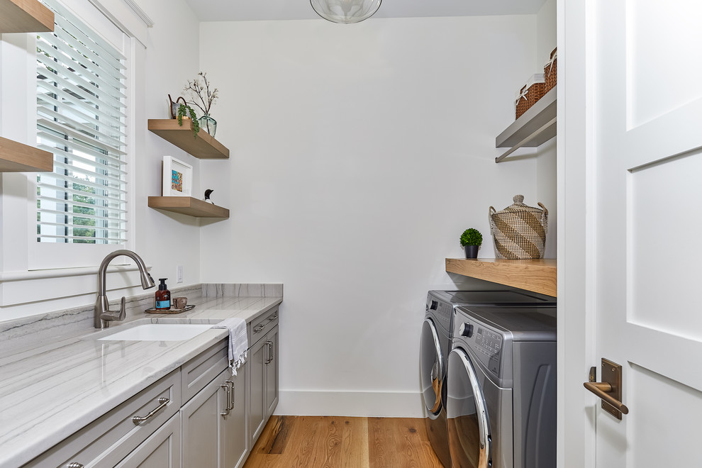Large beach style dedicated laundry room in Charleston with an undermount sink, recessed-panel cabinets, grey cabinets, white walls, light hardwood floors, a side-by-side washer and dryer, grey benchtop and quartzite benchtops.