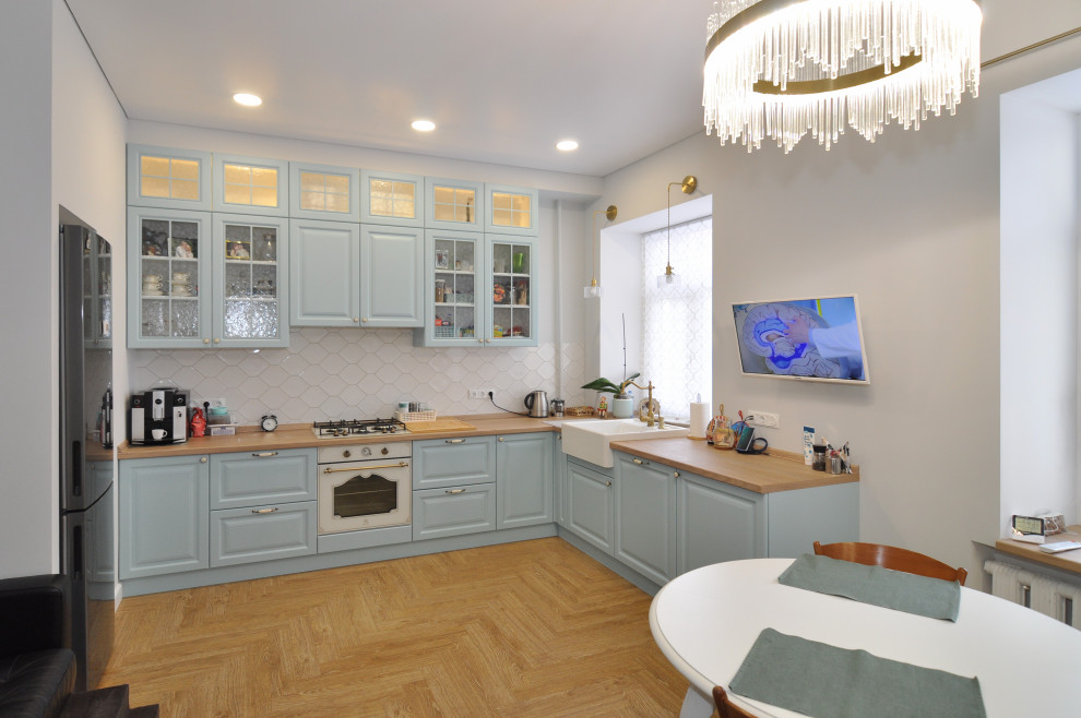 Photo of a mid-sized mediterranean l-shaped eat-in kitchen in Other with a farmhouse sink, raised-panel cabinets, blue cabinets, wood benchtops, white splashback, ceramic splashback, white appliances, medium hardwood floors, no island, beige floor, beige benchtop and recessed.