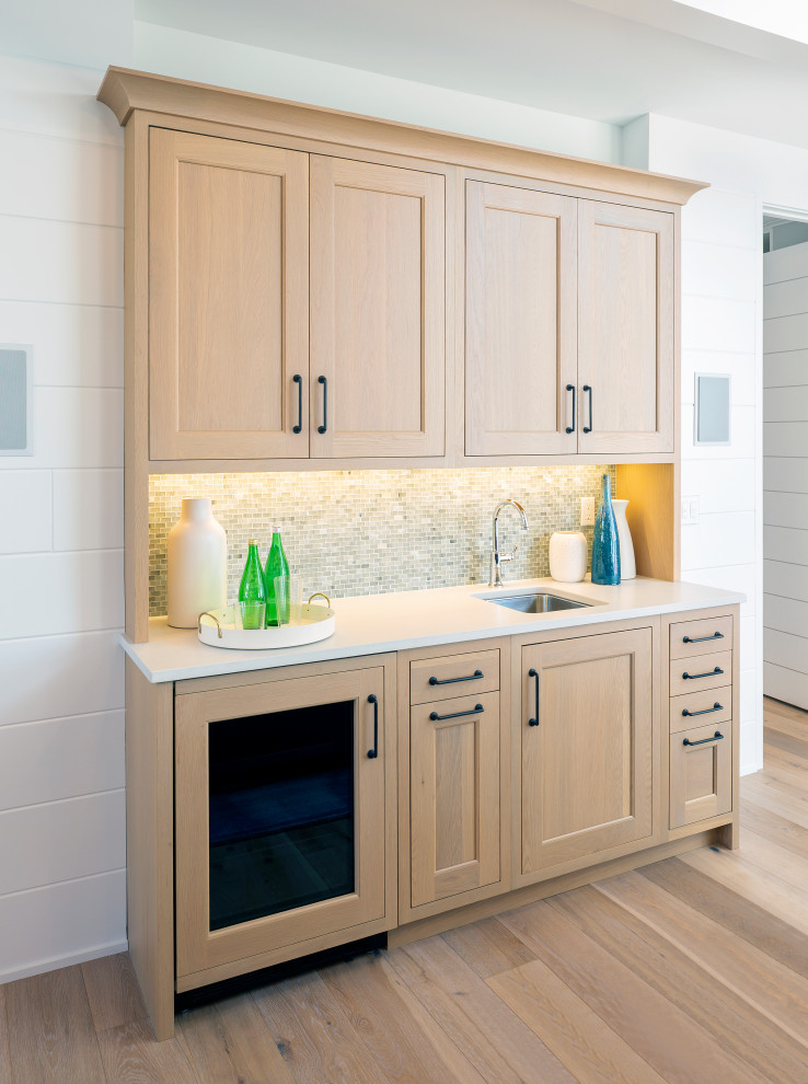 This is an example of a mid-sized transitional single-wall wet bar in Grand Rapids with an undermount sink, recessed-panel cabinets, medium wood cabinets, quartz benchtops, beige splashback, mosaic tile splashback, light hardwood floors and white benchtop.