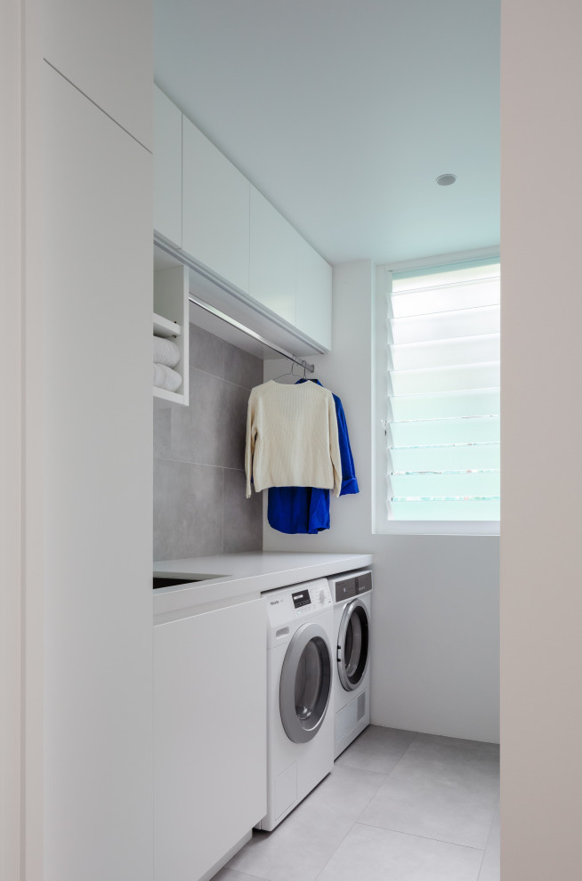 This is an example of a small contemporary single-wall laundry room in Sydney with an undermount sink, quartz benchtops, a side-by-side washer and dryer, flat-panel cabinets, white cabinets, grey splashback, white walls, grey floor and white benchtop.