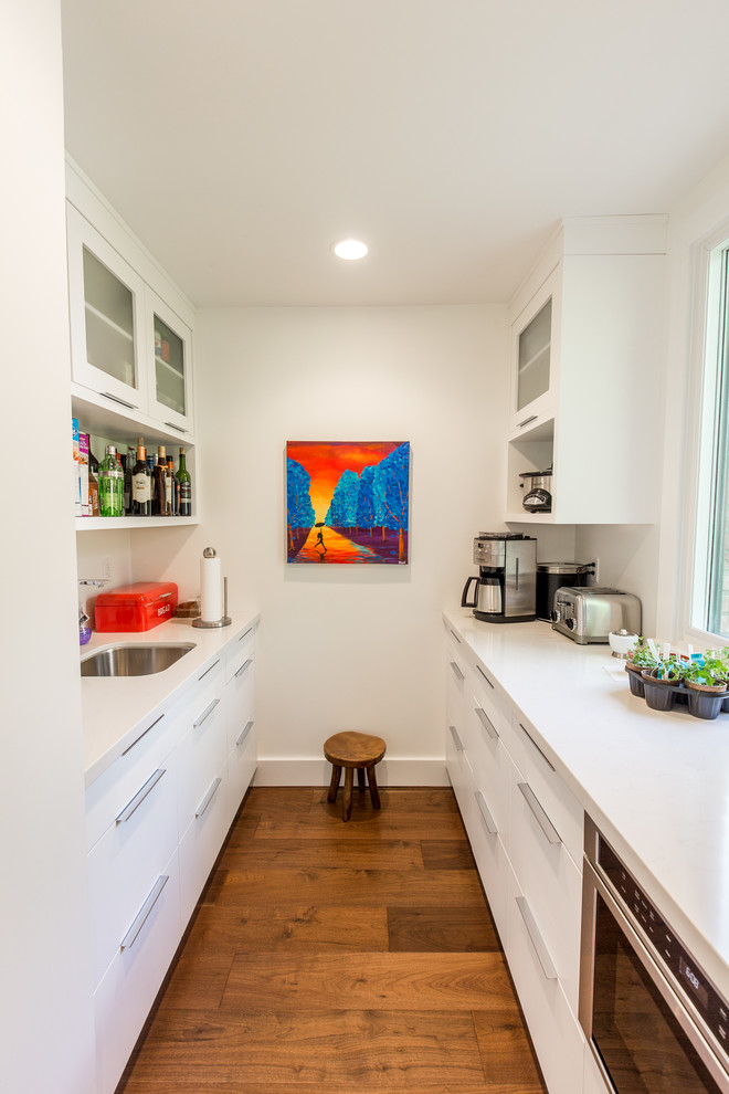 Contemporary galley separate kitchen in Toronto with an undermount sink, flat-panel cabinets, white cabinets, medium hardwood floors, no island, brown floor, solid surface benchtops, white splashback, glass sheet splashback and stainless steel appliances.