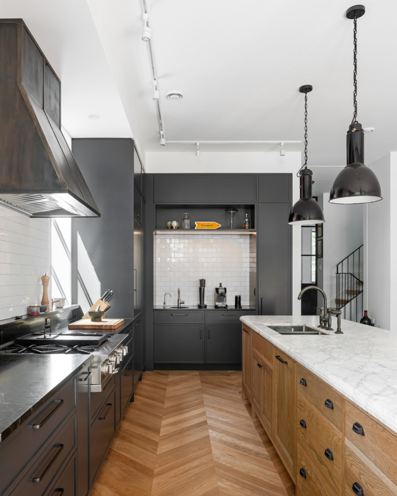 Inspiration for an expansive transitional l-shaped open plan kitchen in Toronto with an undermount sink, recessed-panel cabinets, black cabinets, soapstone benchtops, white splashback, ceramic splashback, stainless steel appliances, light hardwood floors, with island, brown floor and black benchtop.