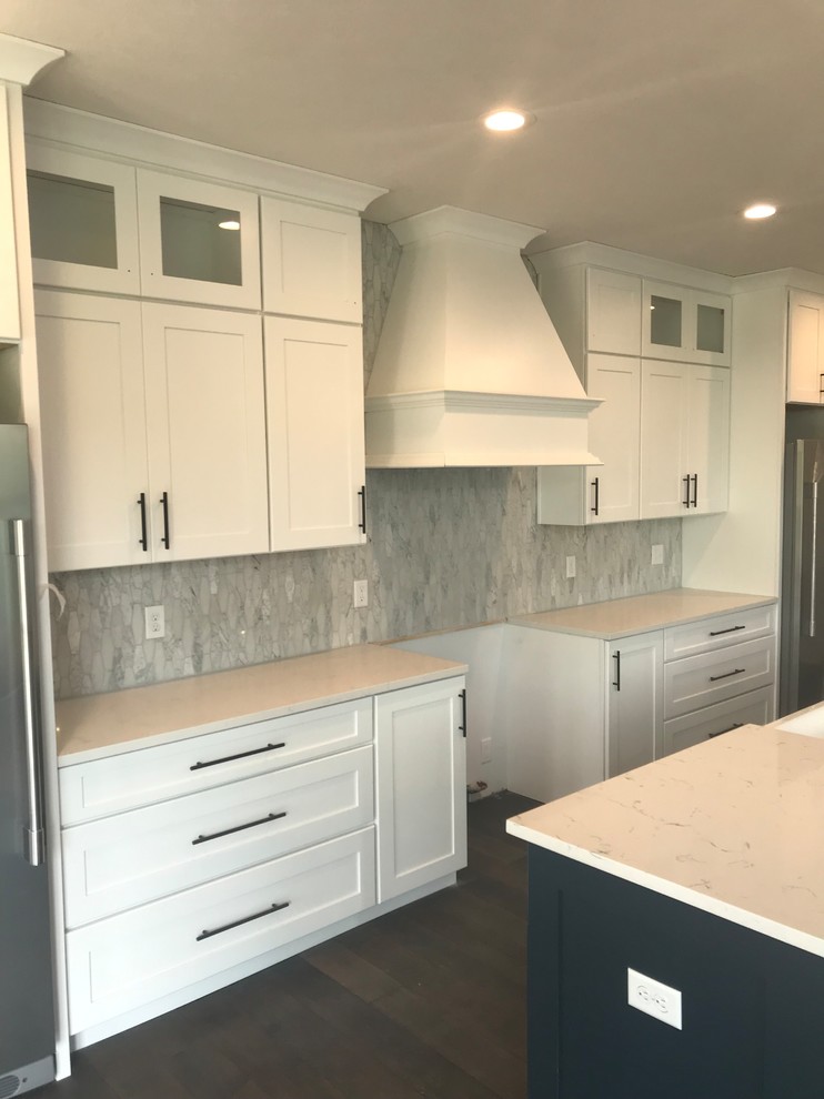 Large transitional single-wall dark wood floor and brown floor open concept kitchen photo with a farmhouse sink, shaker cabinets, white cabinets, solid surface countertops, white backsplash, marble backsplash, stainless steel appliances, an island and white countertops