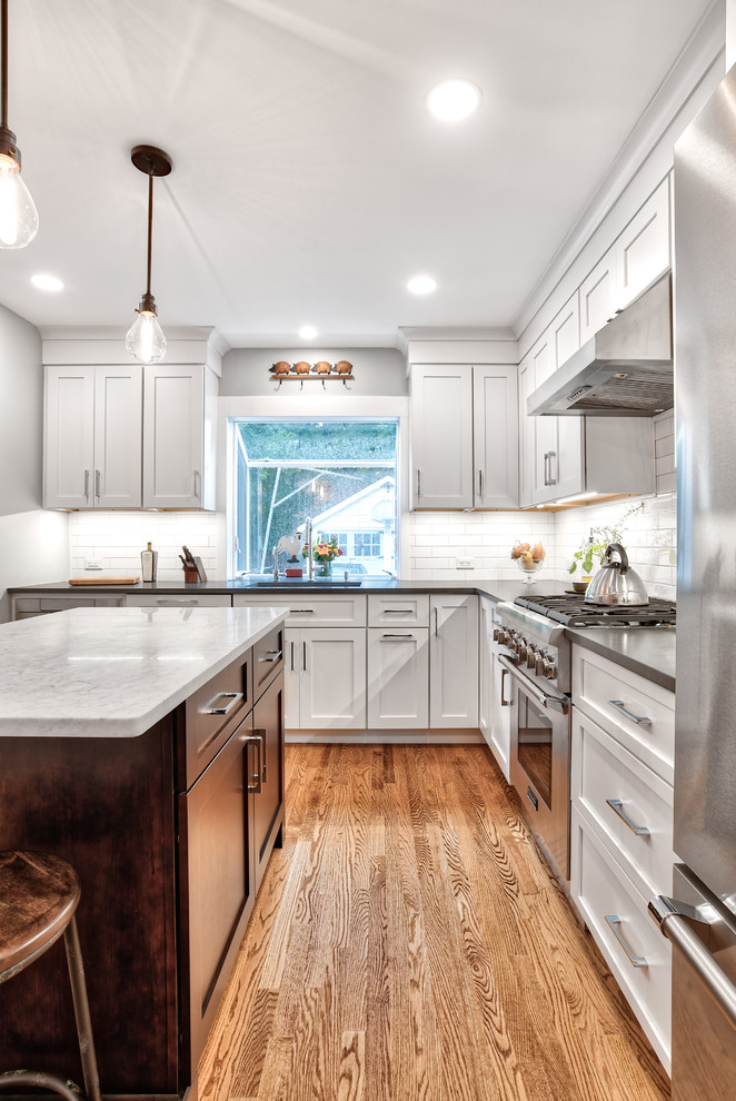 Photo of a mid-sized country l-shaped kitchen pantry in New York with a drop-in sink, beaded inset cabinets, white cabinets, marble benchtops, white splashback, subway tile splashback, stainless steel appliances, painted wood floors, with island, brown floor and black benchtop.