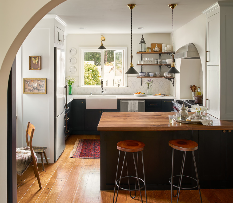 Mid-sized mediterranean u-shaped eat-in kitchen in San Francisco with a farmhouse sink, recessed-panel cabinets, white splashback, porcelain splashback, stainless steel appliances, medium hardwood floors, a peninsula, black cabinets, marble benchtops, brown floor and white benchtop.
