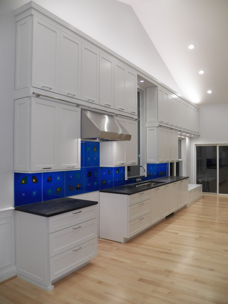 Expansive contemporary galley kitchen pantry in Cincinnati with an undermount sink, shaker cabinets, white cabinets, soapstone benchtops, blue splashback, glass tile splashback, stainless steel appliances, light hardwood floors, with island, black benchtop and vaulted.