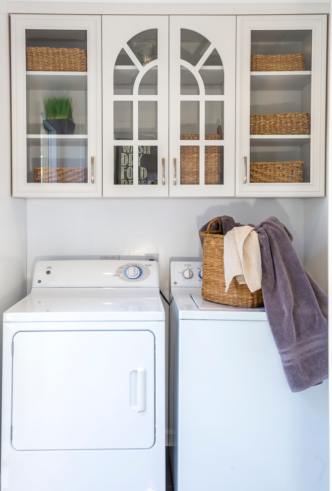 Photo of a small transitional single-wall dedicated laundry room in Los Angeles with glass-front cabinets, white cabinets, white walls, porcelain floors, a side-by-side washer and dryer and grey floor.