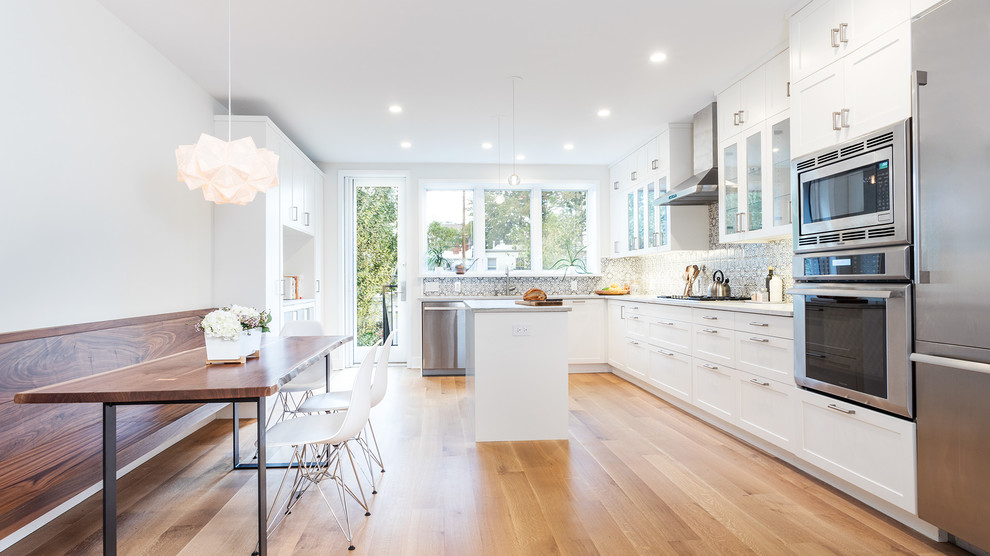 This is an example of a mid-sized transitional l-shaped eat-in kitchen in New York with an undermount sink, shaker cabinets, white cabinets, quartzite benchtops, multi-coloured splashback, porcelain splashback, stainless steel appliances, medium hardwood floors, with island and brown floor.