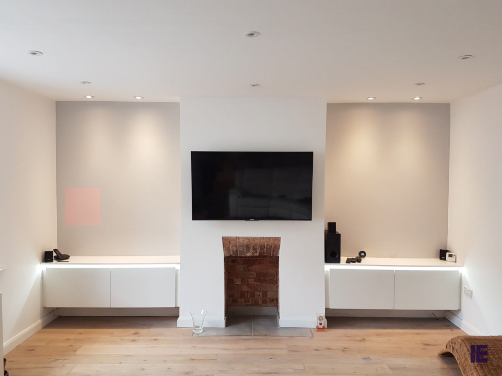 This is an example of a large modern open plan living room in London with white walls, a standard fireplace, a brick fireplace surround, a wall mounted tv and feature lighting.