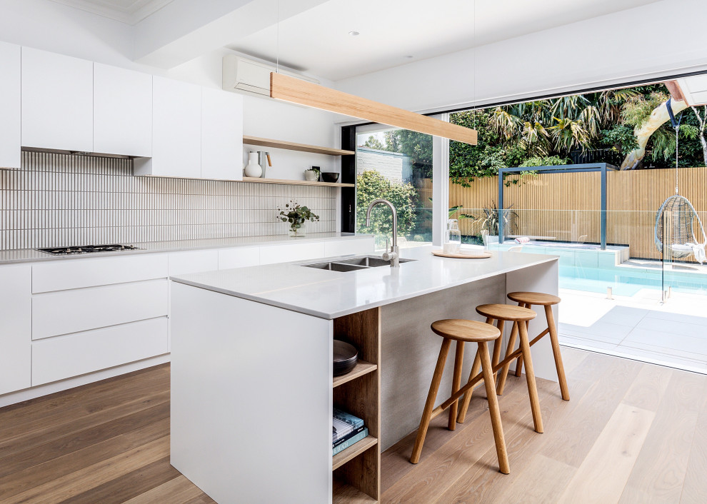 Contemporary galley kitchen in Sydney with an undermount sink, flat-panel cabinets, white cabinets, white splashback, matchstick tile splashback, medium hardwood floors, with island, brown floor and white benchtop.