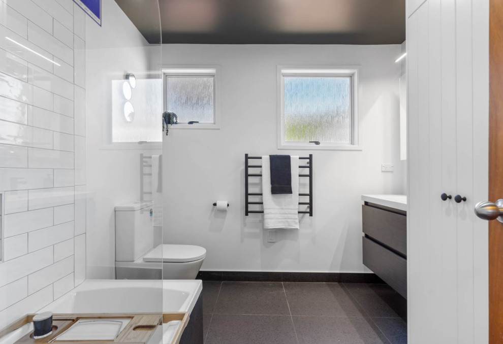 Small contemporary bathroom in Auckland with flat-panel cabinets, dark wood cabinets, a drop-in tub, a shower/bathtub combo, a one-piece toilet, white tile, ceramic tile, white walls, porcelain floors, a drop-in sink, solid surface benchtops, grey floor, a hinged shower door, white benchtops, a single vanity and a floating vanity.