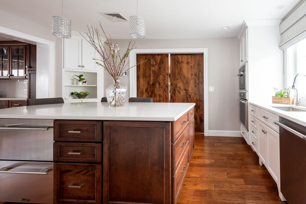Large transitional l-shaped medium tone wood floor and brown floor eat-in kitchen photo in Boston with an undermount sink, shaker cabinets, white cabinets, quartz countertops, white backsplash, subway tile backsplash, stainless steel appliances, an island and white countertops