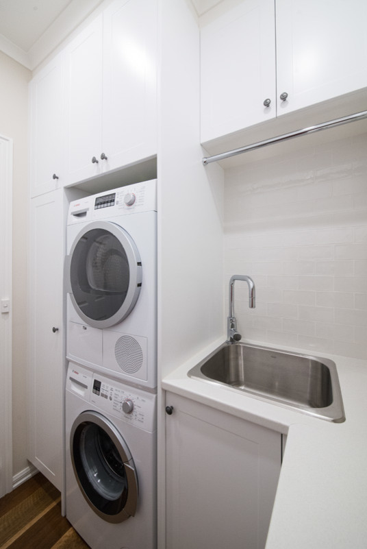 Inspiration for a mid-sized transitional l-shaped dedicated laundry room in Melbourne with an undermount sink, shaker cabinets, white cabinets, granite benchtops, white walls, medium hardwood floors, a stacked washer and dryer and brown floor.
