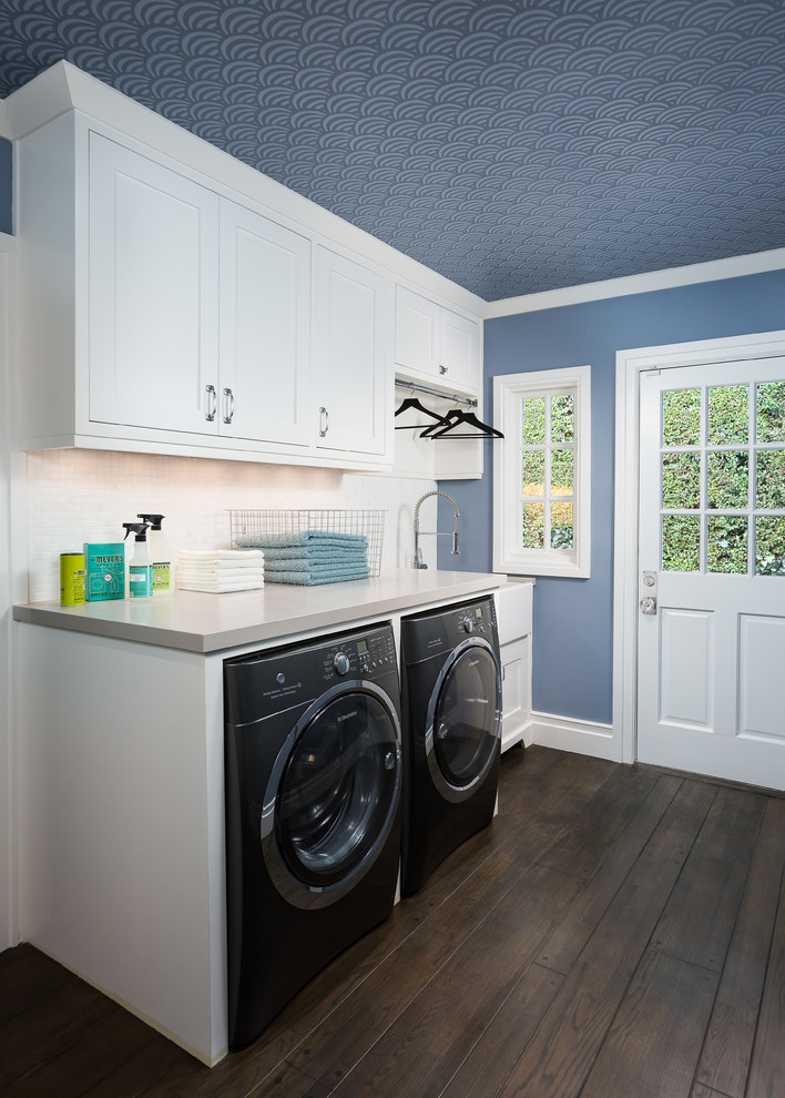 This is an example of a mid-sized transitional single-wall dedicated laundry room in Los Angeles with a farmhouse sink, shaker cabinets, white cabinets, quartz benchtops, blue walls, dark hardwood floors and a side-by-side washer and dryer.