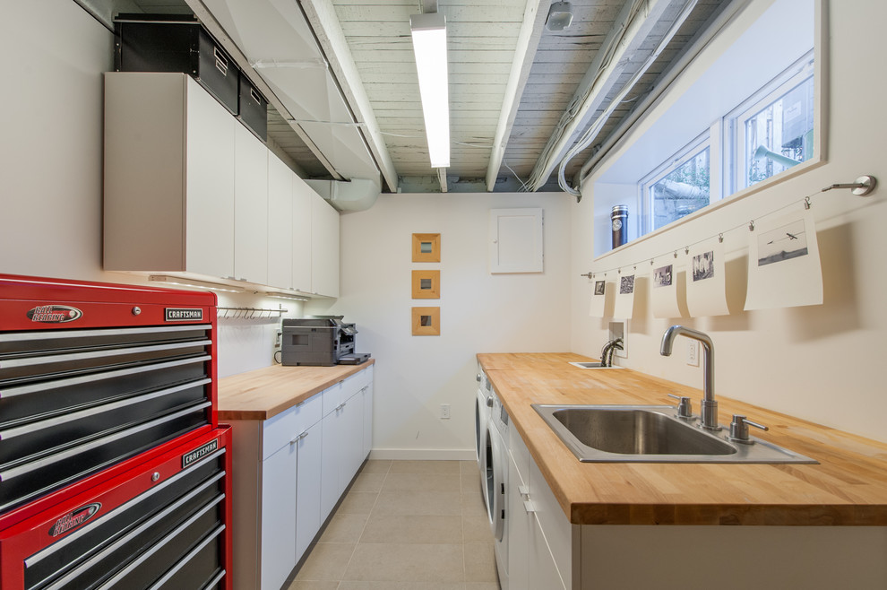 This is an example of a mid-sized industrial galley utility room in DC Metro with a drop-in sink, flat-panel cabinets, white cabinets, wood benchtops, white walls and a side-by-side washer and dryer.