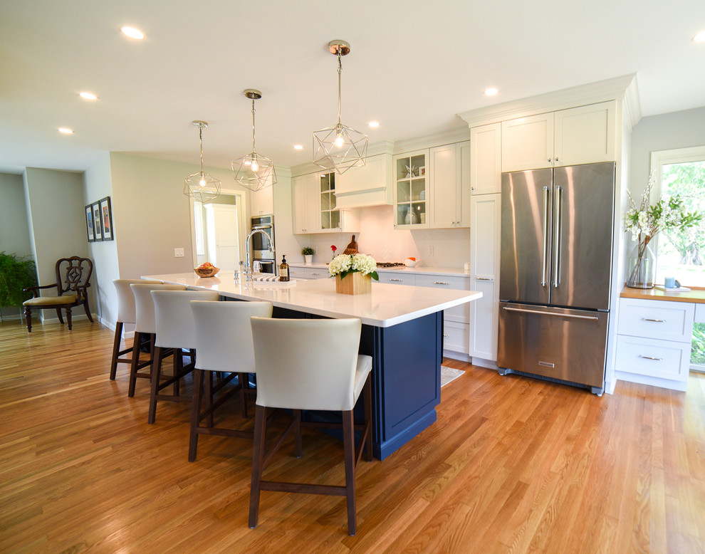Photo of a mid-sized transitional galley open plan kitchen in Philadelphia with a farmhouse sink, shaker cabinets, white cabinets, quartz benchtops, white splashback, ceramic splashback, stainless steel appliances, medium hardwood floors, with island, brown floor and white benchtop.