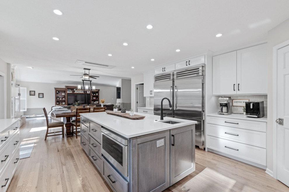 Example of a large transitional u-shaped light wood floor and multicolored floor eat-in kitchen design with a single-bowl sink, shaker cabinets, white cabinets, quartz countertops, white backsplash, quartz backsplash, stainless steel appliances, an island and white countertops