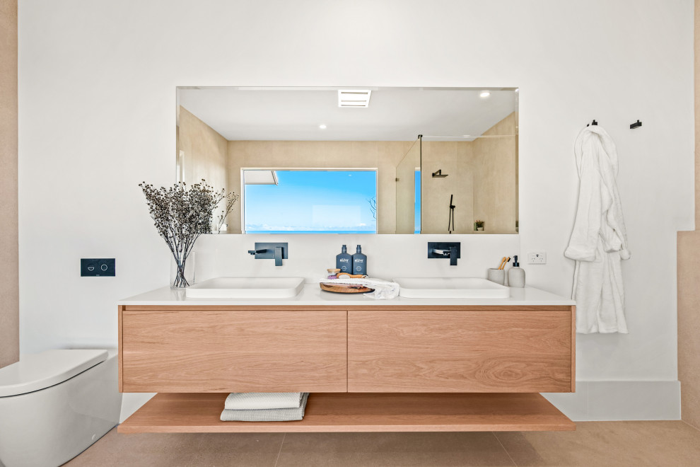 セントラルコーストにあるラグジュアリーな広いビーチスタイルのおしゃれなバスルーム (浴槽なし) (淡色木目調キャビネット、ベージュのタイル、オーバーカウンターシンク、白い洗面カウンター、洗面台2つ、フローティング洗面台、フラットパネル扉のキャビネット、壁掛け式トイレ、白い壁、ベージュの床) の写真