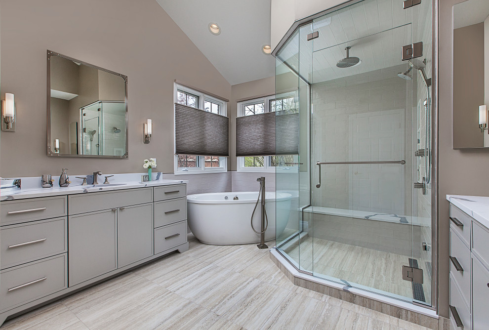 This is an example of a large transitional master bathroom in Detroit with flat-panel cabinets, grey cabinets, a freestanding tub, a corner shower, a bidet, gray tile, ceramic tile, beige walls, ceramic floors, an undermount sink, engineered quartz benchtops, beige floor, a hinged shower door, white benchtops, a shower seat, a double vanity, a built-in vanity and vaulted.