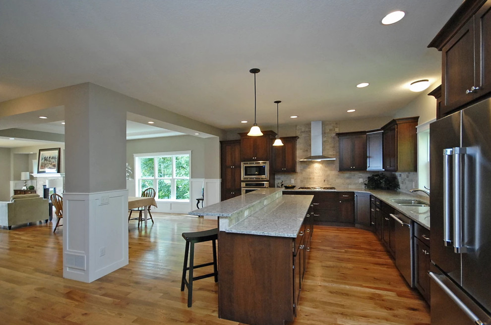 Mid-sized arts and crafts l-shaped open plan kitchen in Minneapolis with a double-bowl sink, recessed-panel cabinets, dark wood cabinets, granite benchtops, grey splashback, porcelain splashback, stainless steel appliances, medium hardwood floors, with island, brown floor and grey benchtop.