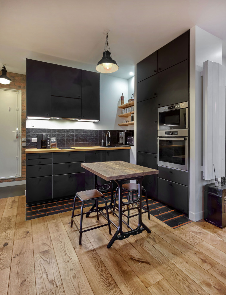 Photo of a small industrial l-shaped eat-in kitchen in Paris with black cabinets, wood benchtops, medium hardwood floors, a double-bowl sink, black splashback, panelled appliances and black floor.